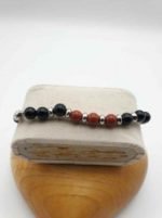 A BRACCIALE DA UOMO CON ONICE NERO, DIASPRO ROSSO E CORDINO CUOIO NERO caratterizzato da perline rosse sopra una scatola di legno.