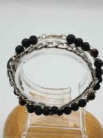 A BRACCIALE UOMO CON PIETRA LAVICA, OCCHIO DI TIGRE E CATENA IN ACCIAIO con perle di lava nera su supporto in legno, con elementi occhio di tigre in acciaio.