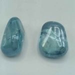 Two AQUA AURA AZZURRA TUMBLED QUALITY A gemstones on a white surface.