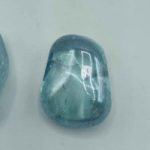 Two Aqua Aura Azzurra Tumbled Quality A stones on a clear surface.